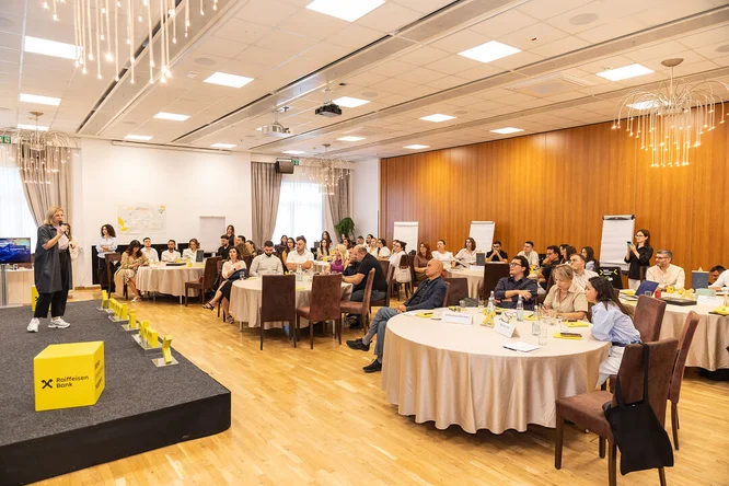 Një audiencë e madhe është ulur në një sallë konferencash duke dëgjuar një folës në skenë. Prezantuesi qëndron përpara një ekrani të madh të verdhë që shfaq tekstin "Të premtet për inovacion" me grafika shumëngjyrëshe. Pjesëmarrësit janë ulur në rreshta karrigesh, me fytyrë nga skena dhe duke i kushtuar vëmendje fjalimit. Ambienti duket profesional dhe modern, me dritat e tavanit dhe pajisjet e prezantimit të dukshme.