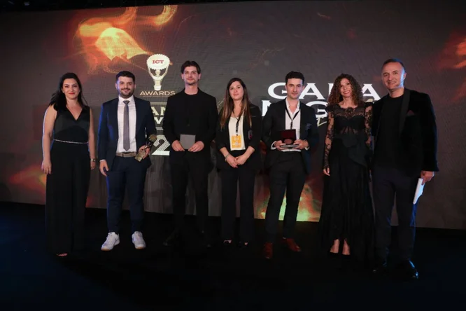A group of seven adults stand on a stage during an awards gala event. Several people hold trophies or plaques, suggesting they are award winners. The backdrop features abstract warm-toned visuals and event text, creating a formal and celebratory atmosphere. The attendees are dressed in elegant evening and business attire under professional stage lighting.