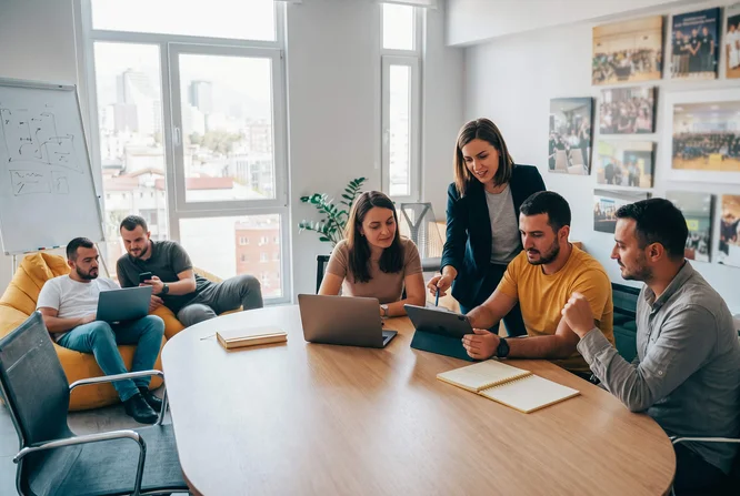 Një grup profesionistësh të rinj bashkëpunojnë rreth një tavoline të madhe prej druri në një zyrë të ndritshme dhe moderne. Disa njerëz përdorin laptopë dhe tableta, ndërsa një grua qëndron dhe tregon me gisht nga një ekran, duke drejtuar diskutimin. Kolegë të tjerë punojnë së bashku në sfond në një divan pranë dritareve të mëdha, me ndërtesat e qytetit të dukshme nga jashtë. Fotot e kornizuara në mur dhe ulëset e rastësishme krijojnë një atmosferë pune bashkëkohore dhe krijuese.
