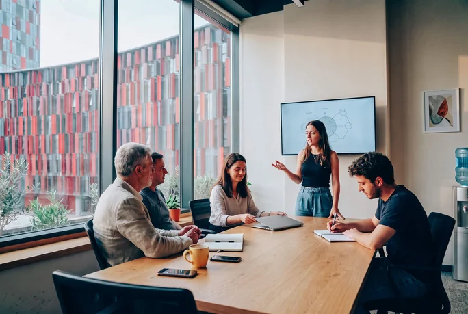A group of five colleagues gather around a wooden conference table in a modern office. One woman stands and speaks to the seated team members, suggesting a presentation or discussion. Large windows reveal a colorful building facade outside, while a wall-mounted screen and water cooler emphasize the workplace setting. Laptops, notebooks, and a smartphone on the table highlight a collaborative business environment.