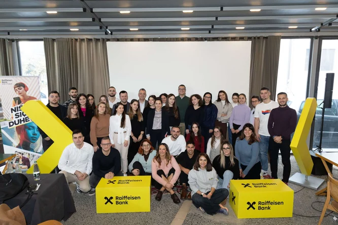 Një grup i madh të rinjsh pozojnë së bashku për një foto në një aktivitet të brendshëm të Raiffeisen Bank. Ata qëndrojnë dhe ulen para një ekrani projeksioni, me panele moderne tavani dhe dritare të mëdha në sfond. Kuba të verdhë të ndezur të Raiffeisen Bank dhe një banderolë vertikale me tekstin "Nje si që duhet" janë të shfaqura në mënyrë të spikatur. Atmosfera duket profesionale, por edhe e rastësishme, duke sugjeruar një punëtori, seminar ose sesion trajnimi.