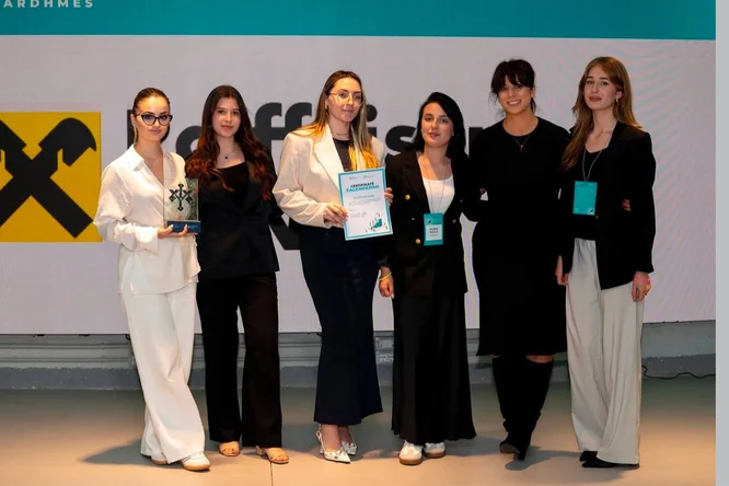 A group of six women stand together on a stage at a professional event. Two of them hold an award trophy and a certificate, suggesting a recognition or achievement ceremony. They are dressed in business and smart casual attire, with event badges visible around their necks. A large screen with a bold yellow and black graphic is displayed in the background, adding to the formal presentation setting.