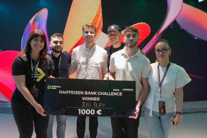 A group of six people stand together on stage holding an oversized check for the Raiffeisen Bank Challenge. The check shows the winner name and a prize amount of 1000 €, dated 04/06/2023. The background features colorful abstract graphics and stage lighting, suggesting a tech or innovation event. The participants wear casual clothing with event badges, smiling and posing for a celebratory photo.