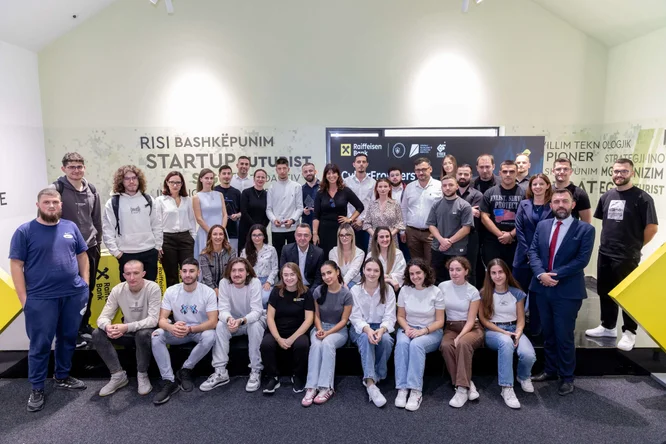 Një grup i madh të rinjsh pozojnë së bashku për një foto ekipore në një hapësirë ​​pune të brendshme për startup-e. Ata janë të vendosur në dy rreshta, me disa të ulur në dysheme dhe të tjerë që qëndrojnë pas tyre. Sfondi paraqet grafika muri me fjalë që lidhen me startup-et dhe inovacionin, së bashku me një panel të zi qendror dhe elementë gjeometrikë të verdhë. Atmosfera duket bashkëpunuese dhe profesionale, me veshje të përditshme dhe disa njerëz me veshje pune.