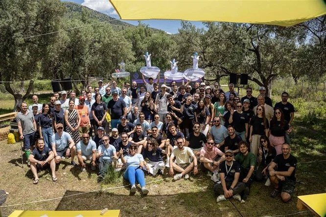 Një grup i madh të rriturish pozojnë së bashku për një foto në një zonë të hapur me bar të rrethuar nga pemë dhe kodra. Skena duket të jetë një aktivitet në natyrë ose një tërheqje ekipore në mot me diell, me struktura hije të verdha që rrethojnë skenën. Njerëzit qëndrojnë dhe ulen afër njëri-tjetrit, duke buzëqeshur dhe duke parë kamerën, duke sugjeruar një atmosferë miqësore dhe festive. Në sfond, dekorimet e fryra dhe pajisjet e aktivitetit janë të dukshme, duke i shtuar ndjesisë festive.