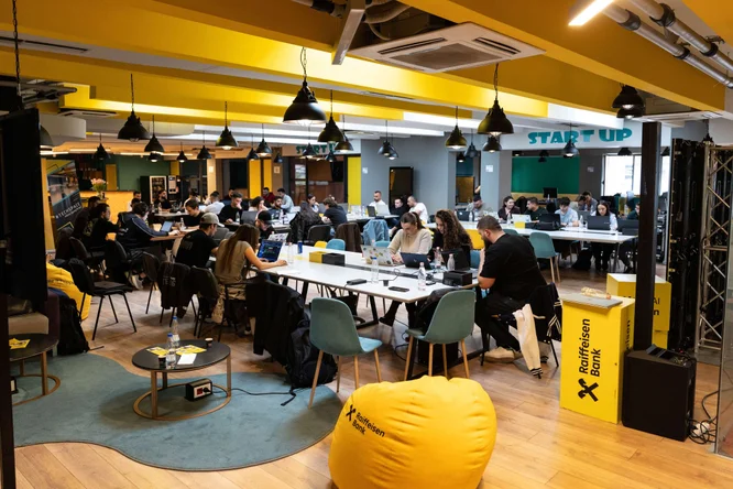 Një grup i madh njerëzish punojnë në tavolina të përbashkëta në një hapësirë ​​moderne pune për startup-e. Pjesa e brendshme përmban detaje të verdha të ndezura, mobilje bashkëkohore dhe ndriçim varës sipër kokës. Teksti i Raiffeisen Bank është i dukshëm në qese të verdha dhe stenda vertikale në plan të parë. Atmosfera duket bashkëpunuese dhe e fokusuar, me laptopë dhe fletore të shpërndara nëpër tavolina.
