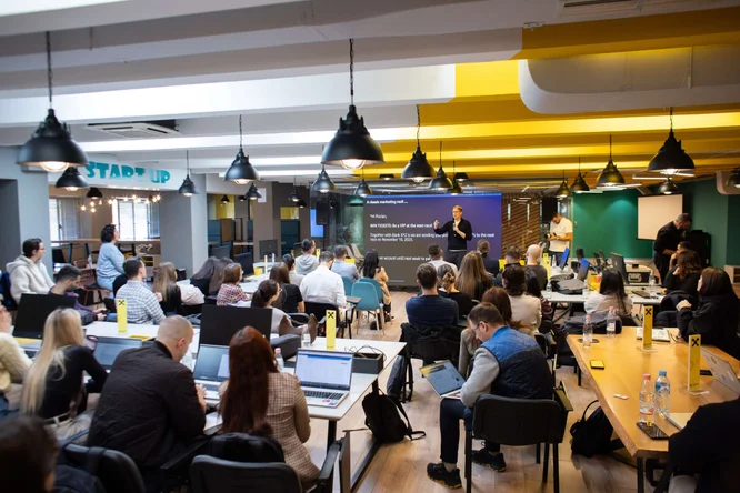 Një grup i madh njerëzish marrin pjesë në një seminar të fokusuar në startup-e në një zyrë moderne bashkëpunuese. Pjesëmarrësit ulen në tavolina të gjata prej druri me laptopë të hapur, ndërsa një prezantues flet para një ekrani të madh. Aksesorët e verdhë të ndezur, llambat e zeza të varura dhe muret prej xhami krijojnë një atmosferë bashkëkohore dhe bashkëpunuese. Dhoma është e mbushur me profesionistë të rinj të angazhuar në të nxënë dhe diskutime.