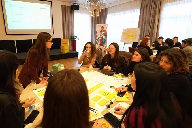 Një grup njerëzish janë ulur rreth një tavoline të angazhuar në një diskutim interaktiv në seminar. Ambienti është një sallë moderne konferencash me dritare të mëdha, perde dhe një llambadar sipër. Letra, kartolina dhe materiale të verdha janë të shpërndara në të gjithë tavolinën, duke sugjeruar aktivitete bashkëpunuese ose shkëmbim idesh. Në sfond, një ekran projektori dhe një tabelë shfletuese janë të dukshme, duke treguar një mjedis formal trajnimi ose seminari.