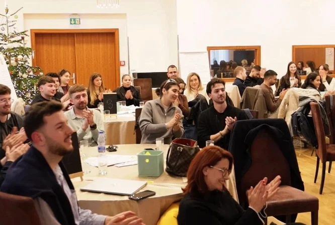 Një grup i madh të rriturish janë ulur rreth tavolinave të rrumbullakëta në një sallë konferencash, duke duartrokitur dhe duke buzëqeshur. Ambienti duket të jetë një seminar profesional ose një takim korporativ me blloqe shënimesh dhe pije mbi tavolina. Një pemë e dekoruar e Krishtlindjeve është e dukshme në sfond, duke sugjeruar një ngjarje të sezonit të festave. Ndriçimi i ngrohtë dhe dekori neutral krijojnë një atmosferë mikpritëse dhe bashkëpunuese.