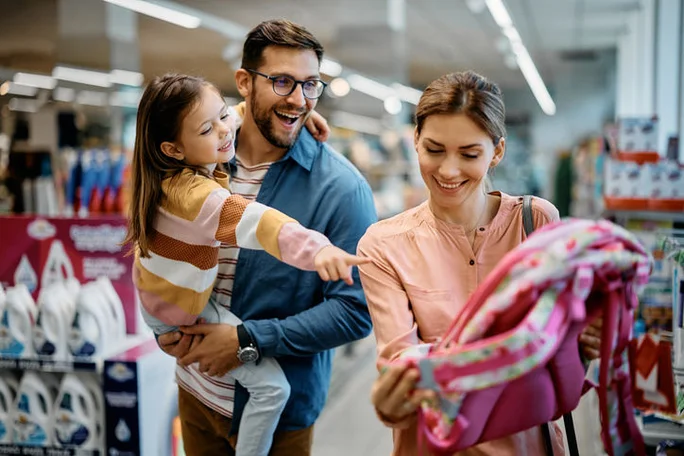 Happy little girl pointing at backpack while buying school supplies with her parents in supermarket.