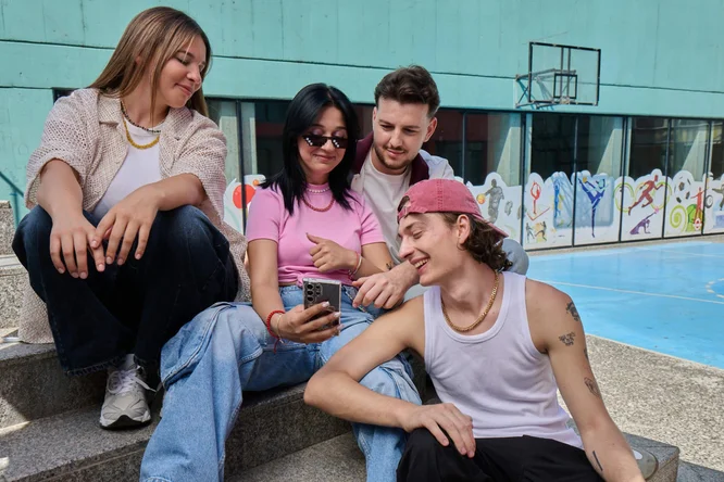 Four young adults sit together on concrete steps near an outdoor sports court, gathered around a smartphone. One person holds the phone while the others lean in, smiling and chatting. The background shows a blue basketball court and colorful wall art, suggesting a casual urban setting. Their relaxed poses and informal clothing convey a friendly, social atmosphere.