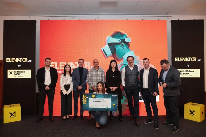 A group of professionals stand on a stage at an Elevator Lab event, with a woman kneeling in front holding a large prize card. The background features a bright orange screen with a person wearing a virtual reality headset and the text "ELEVATOR Lab" clearly visible. Raiffeisen Bank signage appears on both sides of the stage, along with the recognizable black and yellow symbol. The scene suggests a formal award presentation or startup competition in a corporate setting.