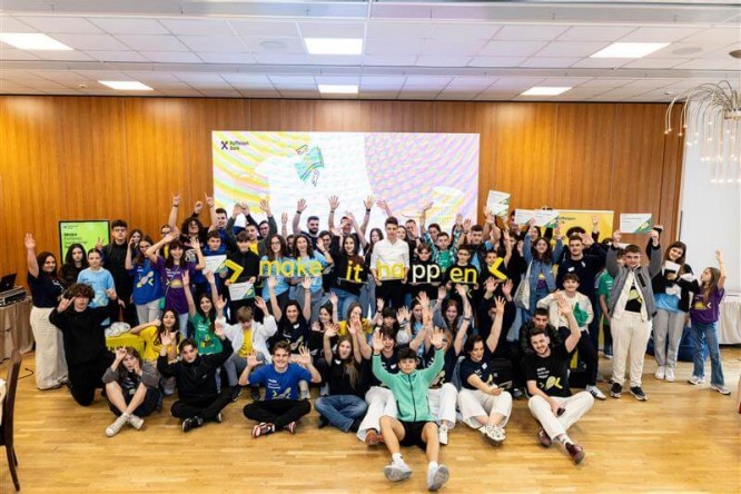 A large group of young people pose together in a bright indoor event space, raising their hands in celebration. The backdrop features colorful graphics and the phrase "make it happen" prominently displayed. Participants wear casual clothing in various vibrant colors and some hold papers or certificates. The wooden wall and polished floor create a warm atmosphere, suggesting a workshop, seminar, or youth program gathering.