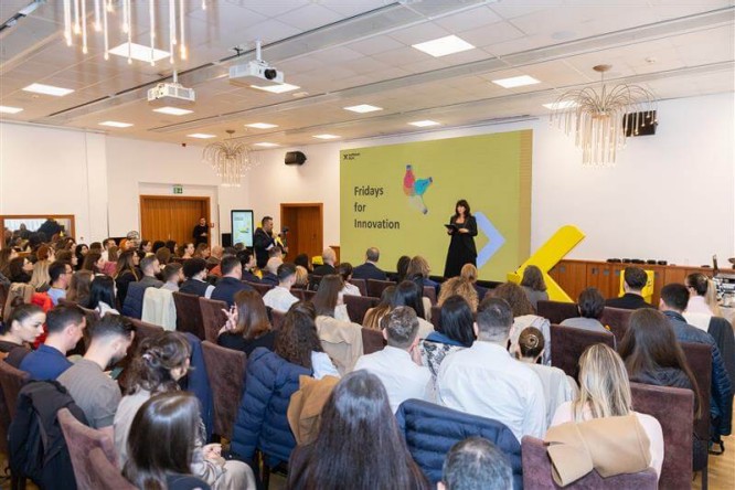 A large audience sits in a conference room listening to a speaker on stage. The presenter stands in front of a big yellow screen displaying the text "Fridays for Innovation" with colorful graphics. Attendees are seated in rows of chairs, facing the stage and paying attention to the talk. The setting appears professional and modern, with ceiling lights and presentation equipment visible.