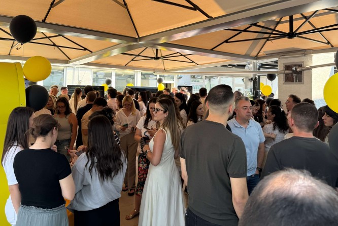 A large group of people gather closely together at an outdoor event beneath beige canopy structures. Many attendees are engaged in conversation, holding drinks and facing different directions. Black and yellow balloons decorate the space, adding a festive touch to the scene. Sunlight filters through the canopy, creating a bright and lively atmosphere.