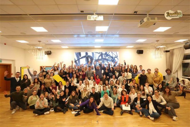 A large group of adults gathers in a spacious indoor conference room, smiling and waving toward the camera. They are seated and standing in several rows on a wooden floor, suggesting a workshop or team event. The room is warmly lit with ceiling lights, and a large artwork or screen is visible at the back wall. Tables, chairs, and presentation boards appear around the edges, reinforcing a collaborative, professional setting.